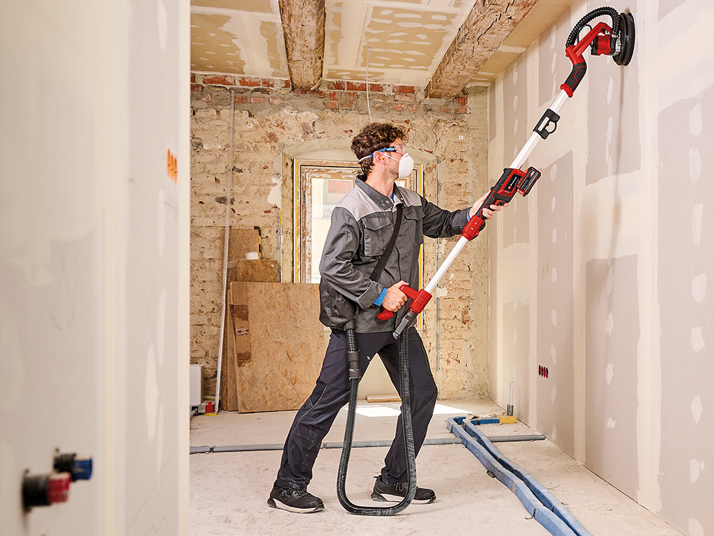 A man sanding drywall with an Einhell cordless drywall sander, its dust bag hanging over his shoulder.