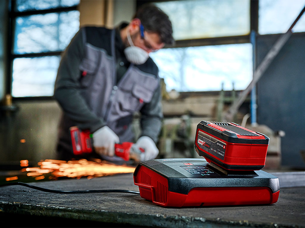 in the front: a Einhell battery charger with a battery | in the back: a man working with a cordless tool