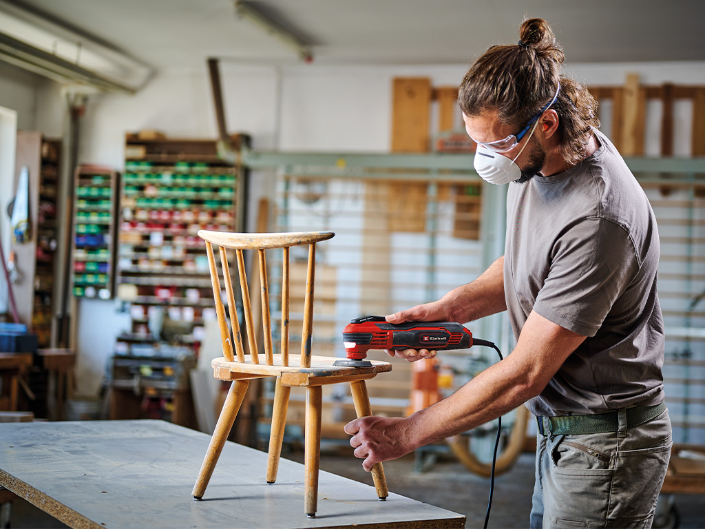 A man sands the frame of a wooden door with a Einhell cordless multitool, wearing safety goggles and a mask.